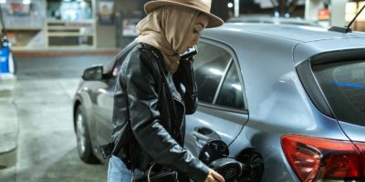 A woman in a hijab and hat refuels her car at a gas station, using a fuel pump.